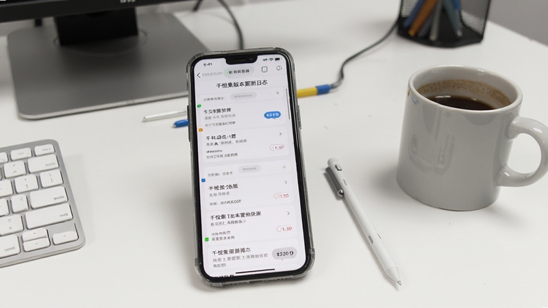 千悦集版本更新日志页面示意图,画面主体为手机屏幕显示更新内容列表,背景为纯白色工作台面,旁边放置一支触控笔和一杯咖啡,营造日常更新维护的场景氛围,适配Boogu模型生成。