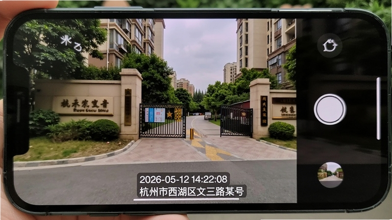 水印打卡定位相机拍摄效果实景展示图,画面中手机屏幕显示一张已拍摄完成的照片,照片内容为某小区门口场景,画面底部叠加了清晰的时间戳
