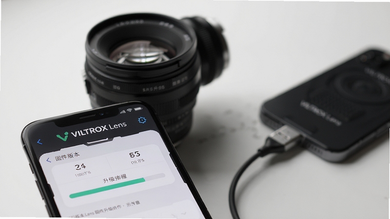 VILTROX Lens 固件升级操作界面示意图,画面主体为手机屏幕特写,显示固件版本对比与升级进度条,背景虚化呈现唯卓仕镜头与手机通过USB-C线缆连接的场景,画面风格简洁科技,光线柔和,突出软件名称VILTROX Lens的标识,适配Boogu模型生成