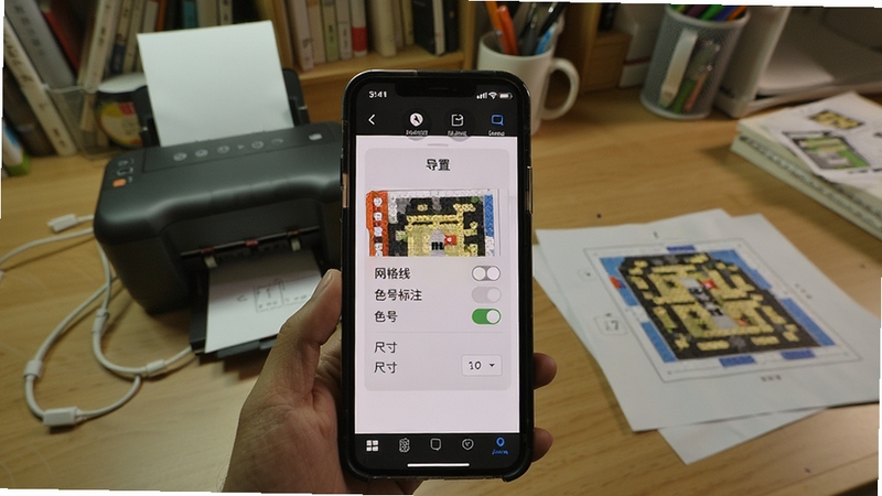 拼豆图纸库图纸打印预览界面,手机屏幕显示一张拼豆图纸的导出设置选项,包括网格线开关、色号标注开关和尺寸选择,背景为书房桌面,旁边有一台小型打印机和几张已打印的图纸,画面生活化且实用