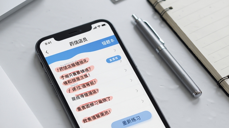 画面主体为手机屏幕特写,显示药信店员版错题本页面,红色标注的错题列表清晰可见,旁边有重新练习按钮。背景区域为浅灰色桌面,放置一支笔和一本笔记本,整体色调清爽,突出学习工具属性。