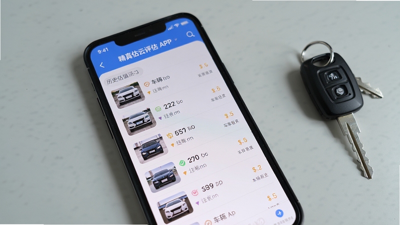 画面主体为精真估云评估APP的历史估值记录列表页面,手机屏幕显示多条车辆估值记录,背景区域为简洁的浅灰色桌面,旁边放着一把车钥匙,整体风格生活化,突出软件日常使用的便利性。