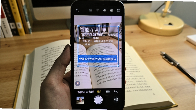 智能万识大师文字识别功能演示,手机拍摄一本翻开的书籍,屏幕上文字被框选并提取为数字文本,界面呈现蓝色高亮效果,背景为书桌场景
