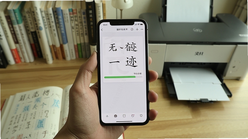 无印有迹移动端操作场景图,画面主体为一只手握持手机,屏幕显示字帖导出进度条,背景区域为书房桌面与打印机,风格生活化写实