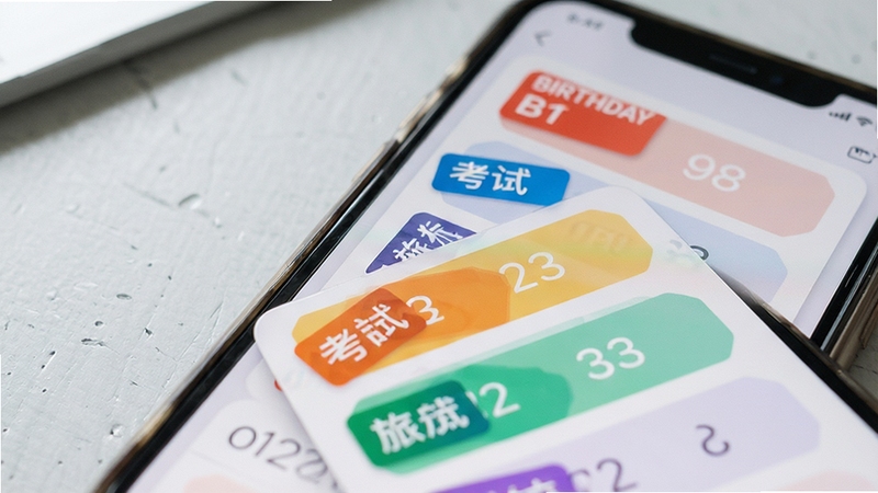 计时倒数日历多事件管理界面特写,手机屏幕内展示不同颜色标签的倒数日列表,包含生日、考试、旅行等分类卡片,背景为浅灰色桌面环境,光线柔和,突出界面层次感