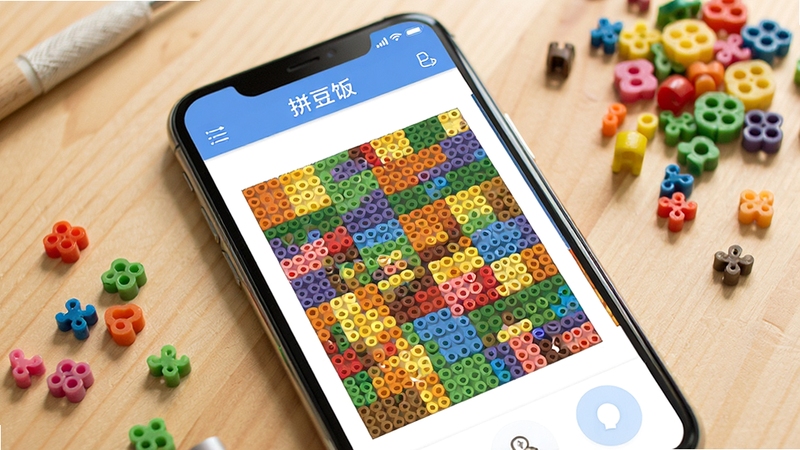 拼豆饭软件主界面展示,画面主体为手机屏幕中显示色彩丰富的拼豆图纸网格,背景为整洁的木纹桌面和散落的彩色拼豆颗粒,整体风格清新明亮,突出拼豆饭软件的核心功能与手工氛围
