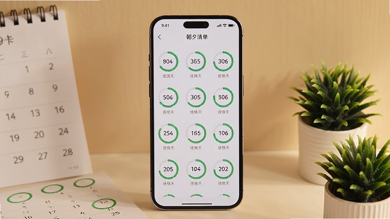 画面中心是一部竖屏手机,屏幕上呈现朝夕清单的习惯打卡页面,多个圆形进度环整齐排列,部分进度环已填满并显示连续天数数字,手机旁边散落着日历页与绿色植物小盆栽,背景为温暖的米色墙面,光线柔和,整体氛围安静自律