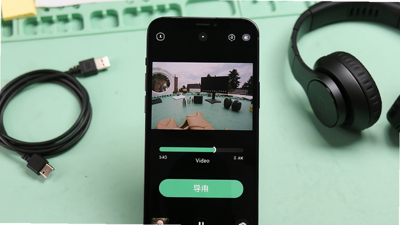 实况live相机免费软件功能展示图,画面中央为手机屏幕显示实况照片转视频的进度条与导出按钮,背景为柔和的浅绿色工作台面,旁边摆放数据线与耳机,突出实用工具氛围