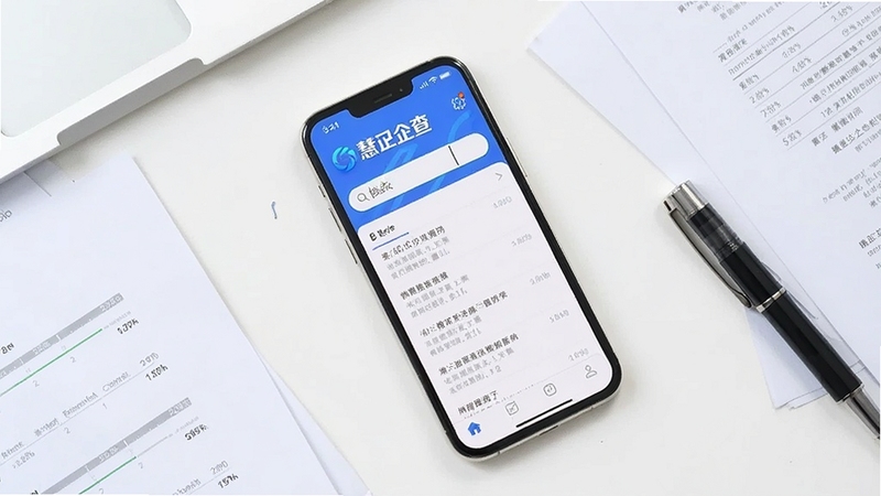 画面采用俯视视角,一部手机平放在白色办公桌上,屏幕显示慧国企的资料查询界面,搜索框内有输入光标,下方列出多条检索结果条目,手机旁边散落几份纸质文件与一支签字笔,整体色调明亮清爽,体现高效查询的工作氛围