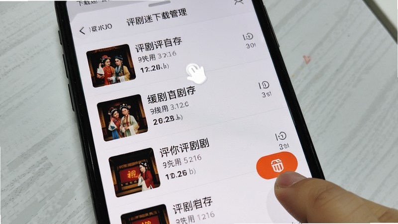 手机屏幕显示评剧迷下载管理界面,列出多个已缓存的评剧剧目及文件大小,旁边有手指点击删除按钮的动作示意,背景为简洁的浅灰色桌面,突出离线缓存管理功能
