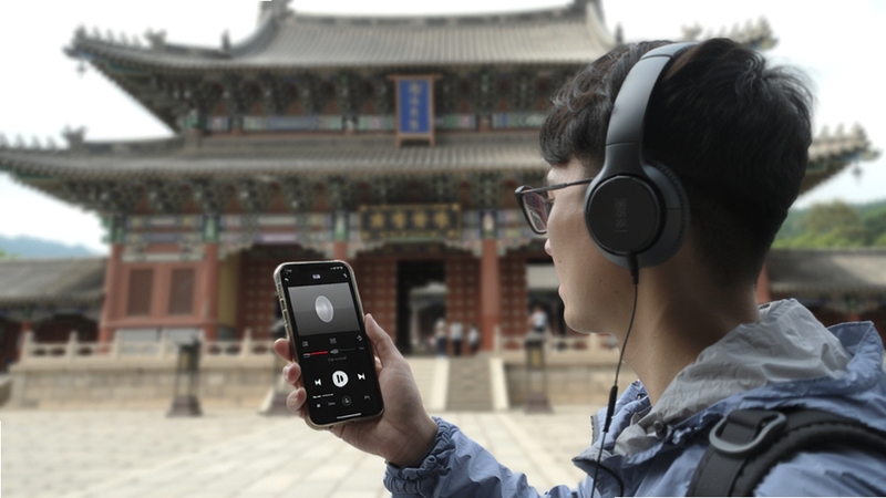 旅途语音解说功能场景,一位游客戴着耳机站在古建筑前,手机屏幕显示音频播放界面,背景虚化,突出人物与手机,风格写实