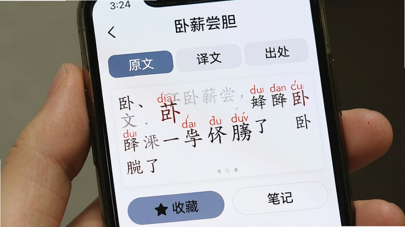 手机界面特写,屏幕中展示一条典故详情,顶部是词条名称“卧薪尝胆”,下方分为“原文”“译文”“出处”三个标签页,当前“原文”页高亮,文字中若干生僻字上方标注了红色拼音,页面底部有“收藏”和“笔记”两个按钮,界面风格与前面一致,整体观感专业且友好。