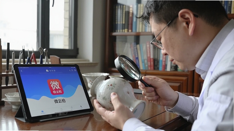 微云拍专家鉴定场景,一位专家手持放大镜仔细查看瓷器,旁边摆放微云拍APP平板界面,背景为工作室书柜,自然光,细节清晰,展示鉴定过程与专业感