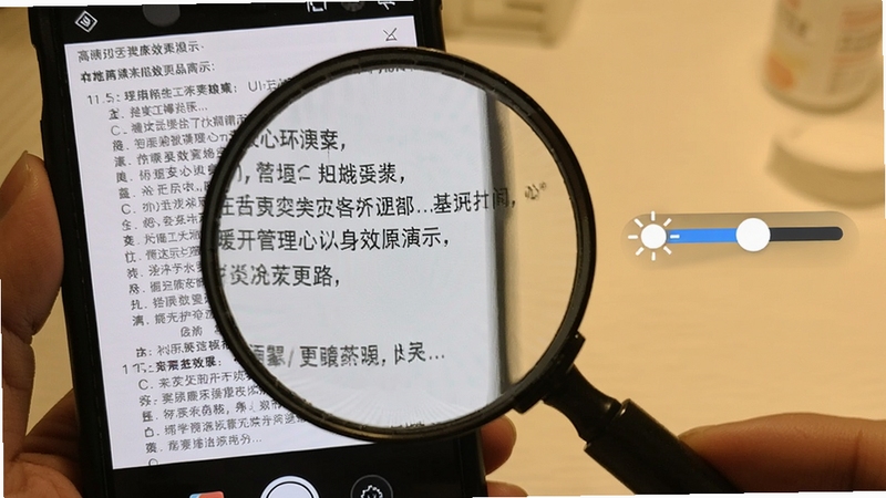暖心放大镜的放大效果演示,手机摄像头对准药品说明书,屏幕上清晰显示出放大的文字,文字边缘锐利,背景光线柔和,旁边配有亮度调节滑块,展示高清放大的细节。