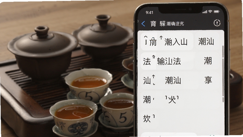 潮语输入法拼音联想功能演示图,手机屏幕上显示输入潮汕拼音后出现多个汉字候选词,背景为潮汕工夫茶具,突出智能联想与准确识别