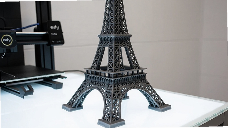 Boogu生成,一张展示eufyMake打印完成的精美3D模型成品的照片,模型是一个复杂的埃菲尔铁塔,细节丰富,表面光滑,摆放于明亮的展示台上,背景是简洁的工作室环境,旁边放置着eufyMake打印机机身一角,画面清晰锐利,突出打印质量与机器质感。