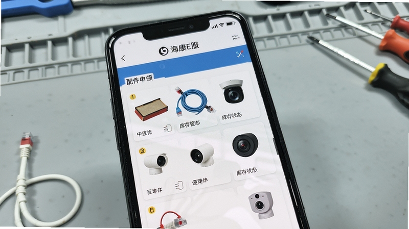 海康E服 配件申领页面 手机屏幕展示配件商城列表与库存状态 画面中有螺丝刀、网线、摄像头等元素 背景为浅灰色工作台 风格写实 突出便捷申领与库存管理