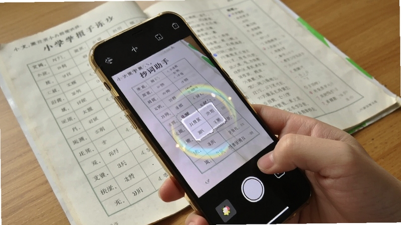 抄词助手拍照识别功能特写,画面主体为一只手持手机对准课本生词表进行拍摄,手机屏幕显示识别中的动态光效,背景是翻开的小学语文课本与木质桌面,光线柔和,突出智能识别与便捷录入的场景