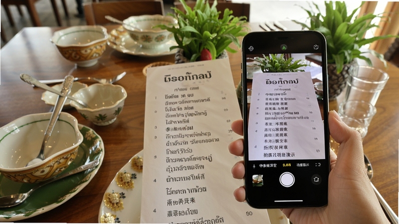 中泰翻译官拍照翻译功能演示,画面主体为手机摄像头对准一份泰文菜单,手机屏幕显示识别后的中文翻译结果,背景为泰国餐厅桌面,有泰式餐具和绿植点缀,画面清晰明亮,适配Boogu模型生成