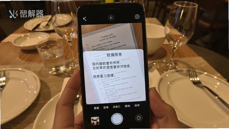 翻译器拍照翻译操作演示图,画面展示手机摄像头对准一份英文菜单,屏幕浮层显示中文译文,背景为餐厅餐桌场景,突出翻译器软件名称与拍照识别界面细节